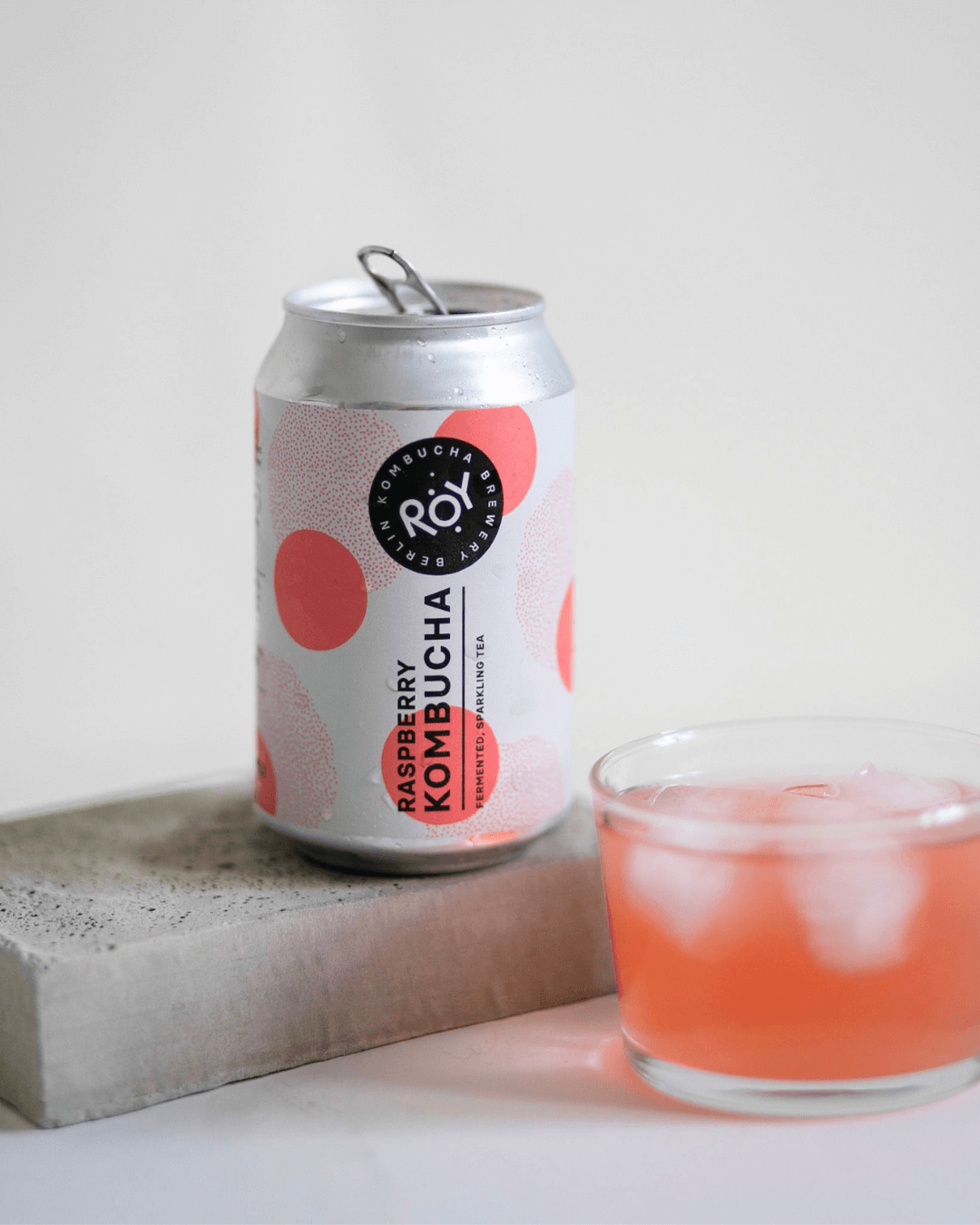 Bio-Himbeer-Kombucha von ROY Kombucha in einer Dose mit rosa Design, daneben ein Glas mit Eiswürfeln.