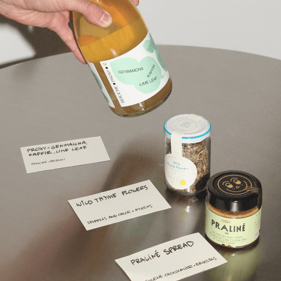 Drei Feinkostprodukte auf einem Stahltisch: ein Getränk von Bouche, ein Glas mit Thymianblüten, ein Glas mit Praliné Nussmus.