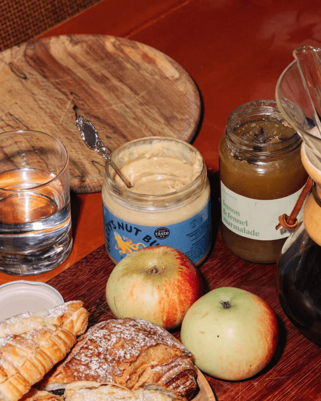 Ein Glas Harry’s Nut Butter Crunchy mit einem Löffel, umgeben von Äpfeln, Croissants, Marmelade auf einem Tisch.