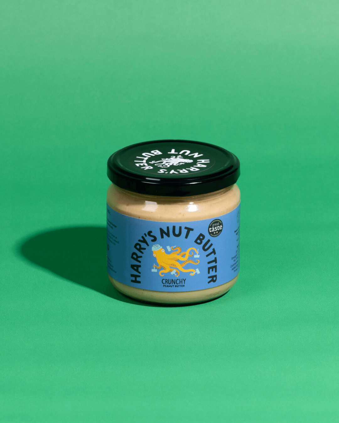 Ein Glas Harry’s Nut Butter Crunchy mit blauem Etikett und schwarzem Deckel, platziert auf einem grünen Hintergrund.