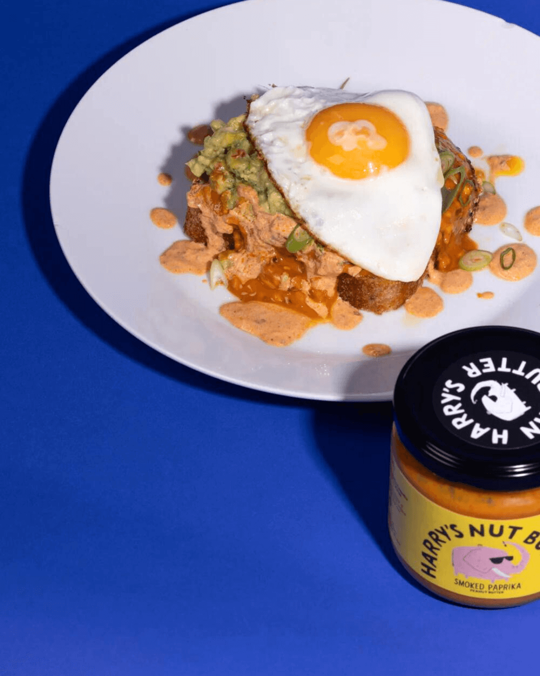 Gericht mit Spiegelei, Guacamole und Soße auf Brot, daneben ein Glas Harry's Erdnussbutter mit geräuchertem Paprika.