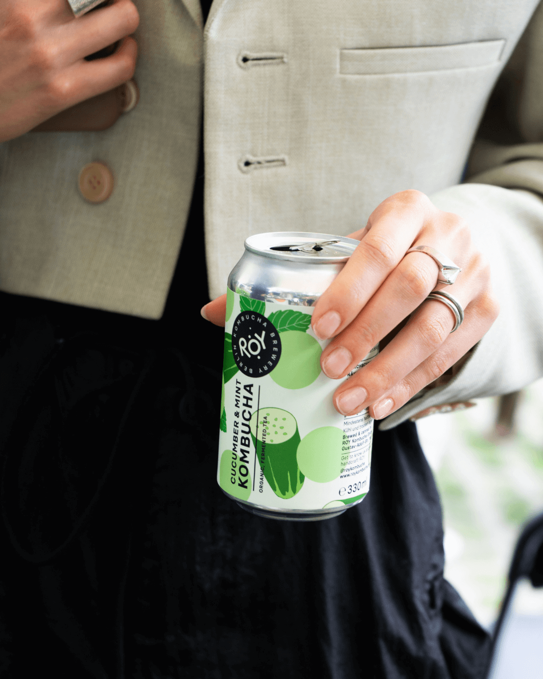 Hand hält eine Dose ROY Kombucha Gurke & Minze, stilvoll in beiger Jacke und schwarzen Stoffen.